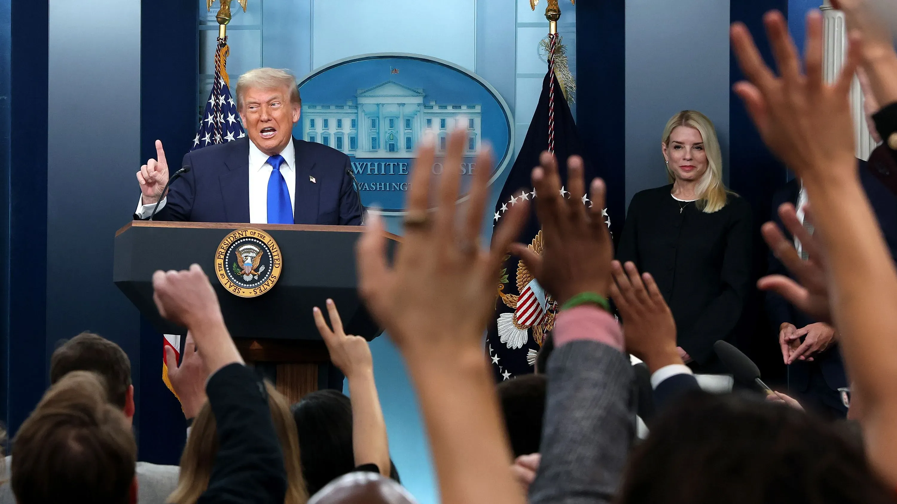 ترامب يتحدث في مؤتمر صحفي بالبيت الأبيض، حيث ترفع مجموعة من الصحفيين أيديهم لطرح الأسئلة، بينما تقف بجانبه مديرة الاتصالات.