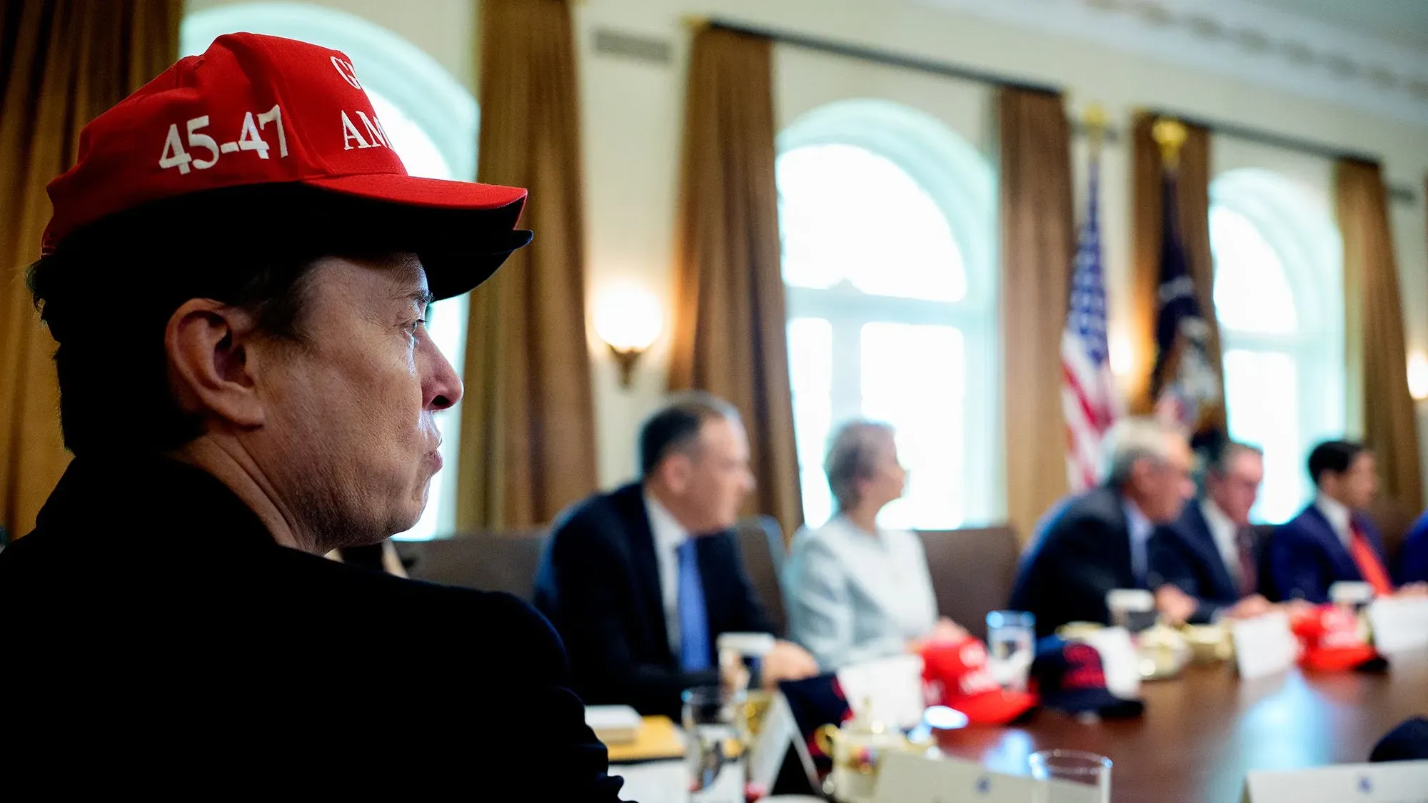 رجل يرتدي قبعة حمراء تحمل الرقمين 45-47، يجلس في اجتماع مع مسؤولين حكوميين في البيت الأبيض، حيث تثار قضايا حول الشفافية الحكومية.