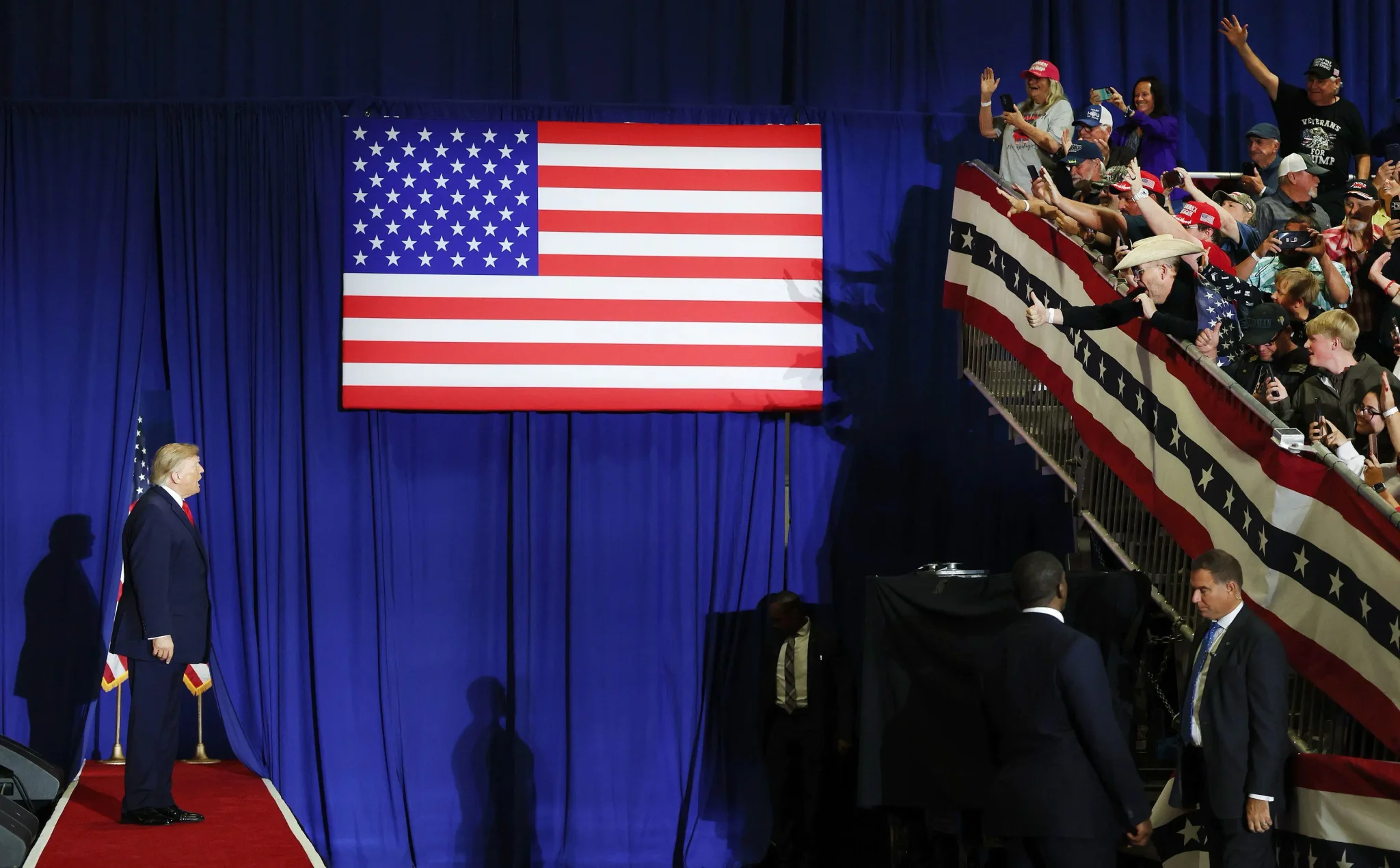 دونالد ترامب يقف على منصة حمراء أمام جمهور متحمس يحمل أعلام الولايات المتحدة في فعالية سياسية داخلية، مع خلفية زرقاء وعلم أمريكي كبير.