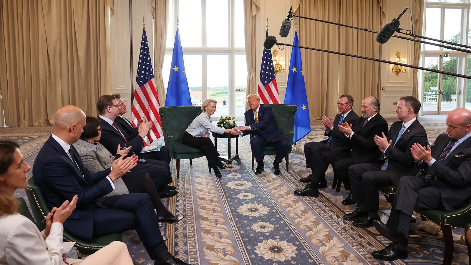 ترامب يتصافح مع مسؤولة من الاتحاد الأوروبي خلال اجتماع تجاري، بينما يجلس المفاوضون ويصفقون في خلفية الحدث.