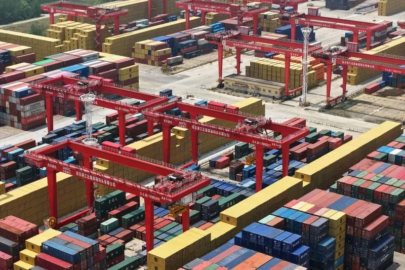 صورة لميناء شحن مزدحم يحتوي على حاويات ملونة وآلات رافعة حمراء، تعكس التوترات التجارية بين الولايات المتحدة والصين.