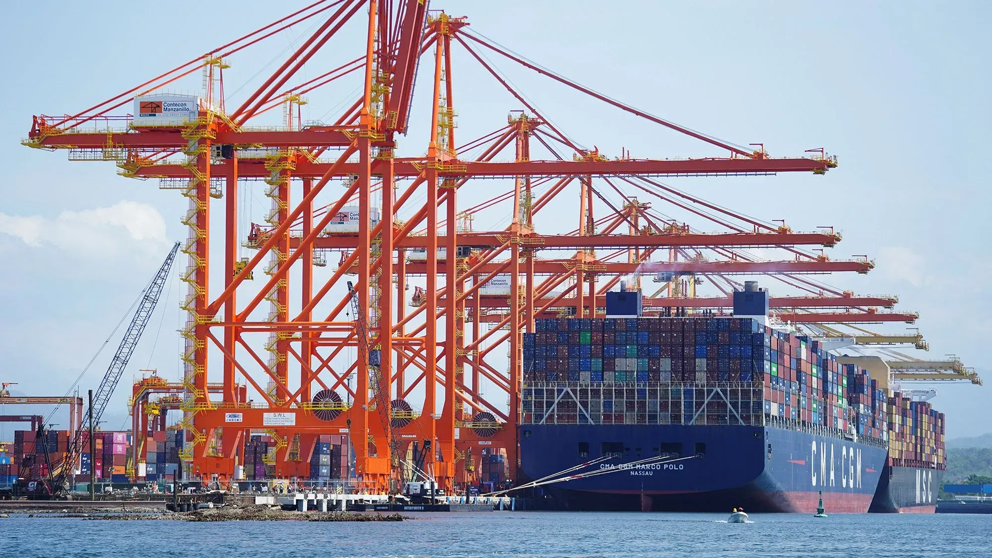 سفينة حاويات كبيرة تخضع للتفريغ في ميناء، مع رافعات حمراء ضخمة وآلاف الحاويات المتنوعة، تعكس التبادل التجاري بين أمريكا وكندا والمكسيك.