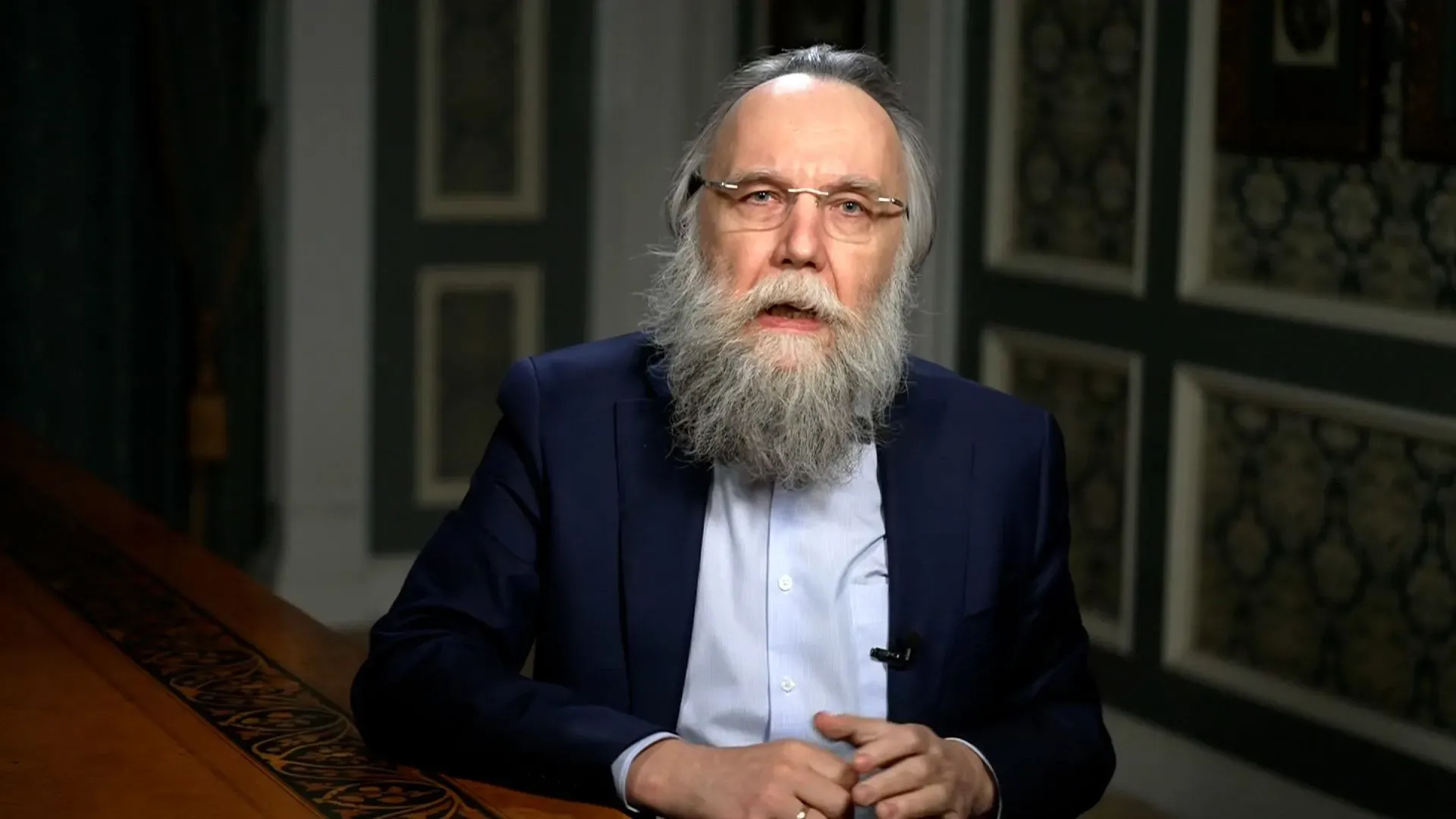 رجل ذو لحية طويلة يرتدي بدلة، يتحدث أمام كاميرا في بيئة داخلية مزينة. تعكس تعابيره الجدية والتفكير العميق.