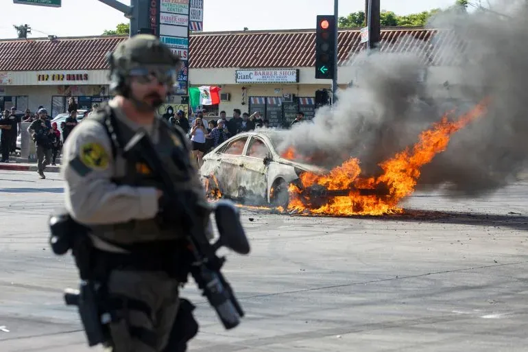 شرطي مسلح يسير بجانب سيارة مشتعلة في احتجاجات لوس أنجلوس، حيث تصاعد الدخان والنيران وسط حشود المتظاهرين.