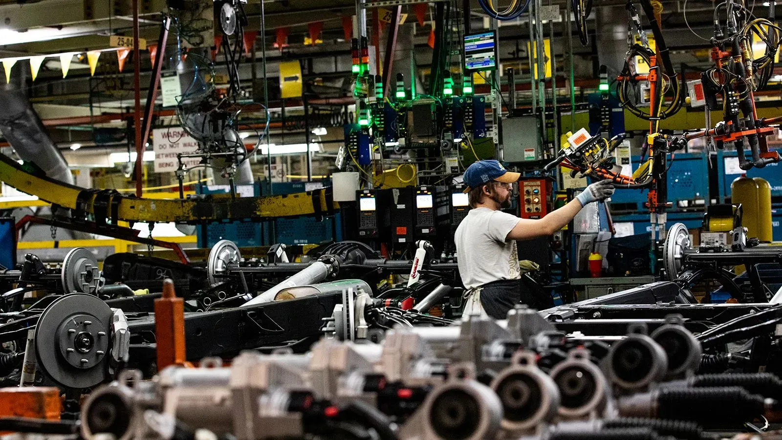 عامل في مصنع سيارات يعمل على تجميع أجزاء المركبات، مع وجود معدات وآلات في الخلفية، يعكس تأثير التعريفات الجمركية على الإنتاج.