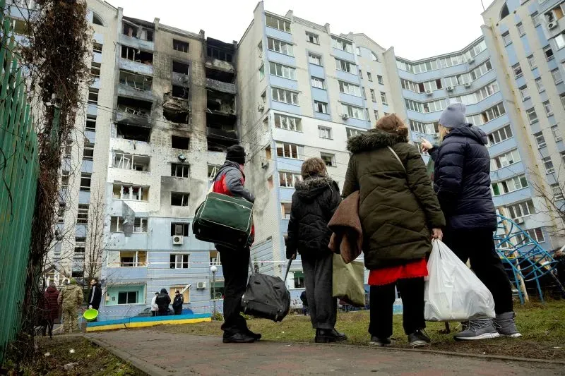 مجموعة من الأشخاص يقفون أمام مبنى سكني متضرر في أوكرانيا، حيث تظهر آثار الدمار على الواجهة، مما يعكس تأثير النزاع المستمر.