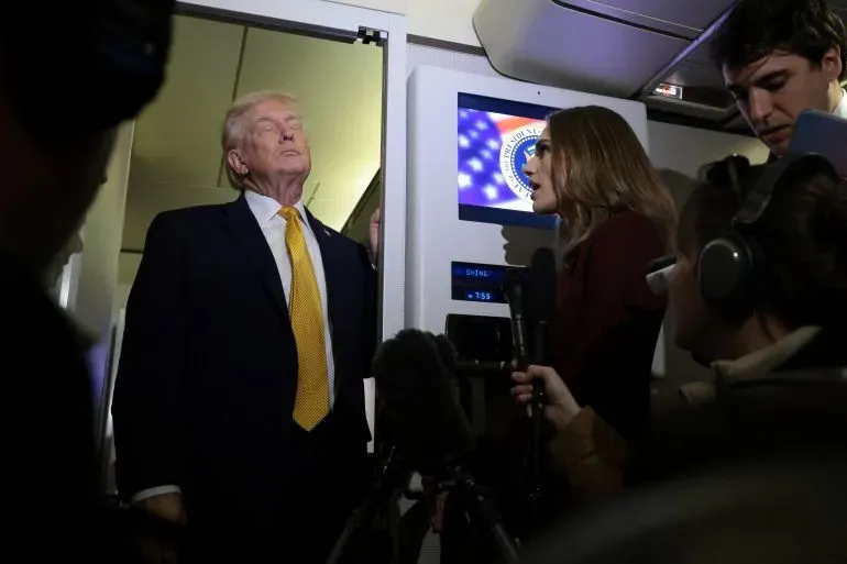 دونالد ترامب يقف أمام باب الطائرة، بينما تتحدث صحفية إليه، محاطًا بعدد من الصحفيين، مع شاشة تعرض شعار الحكومة الأمريكية في الخلفية.