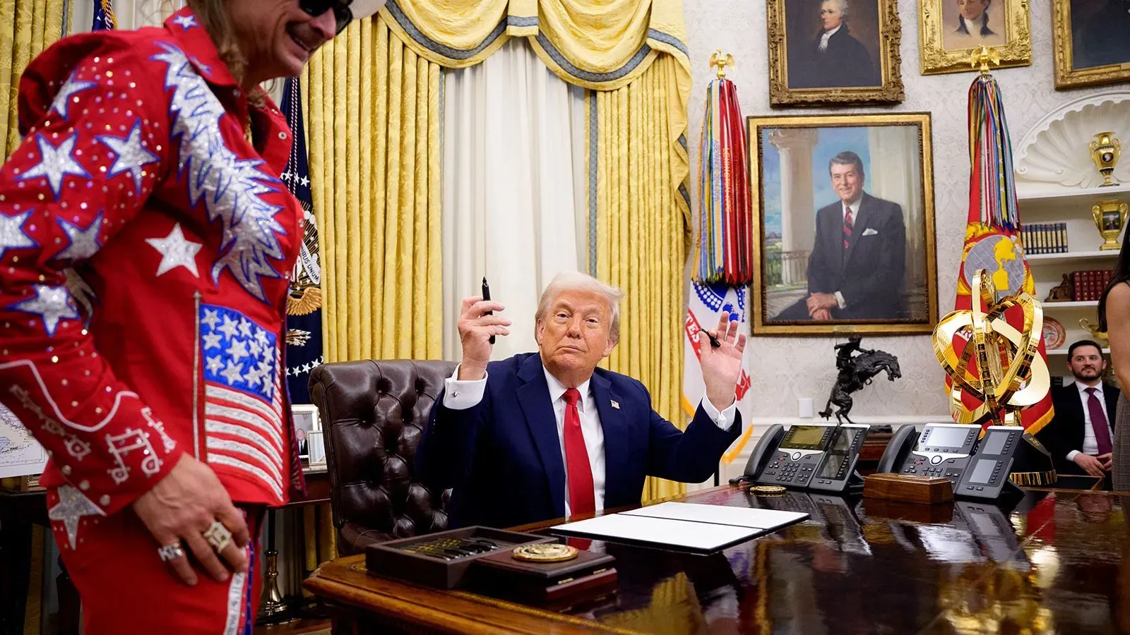 ترامب يجلس في المكتب البيضاوي، محاطًا بكيد روك الذي يرتدي بدلة ملونة، أثناء توقيع قرار بشأن الرسوم الجمركية.