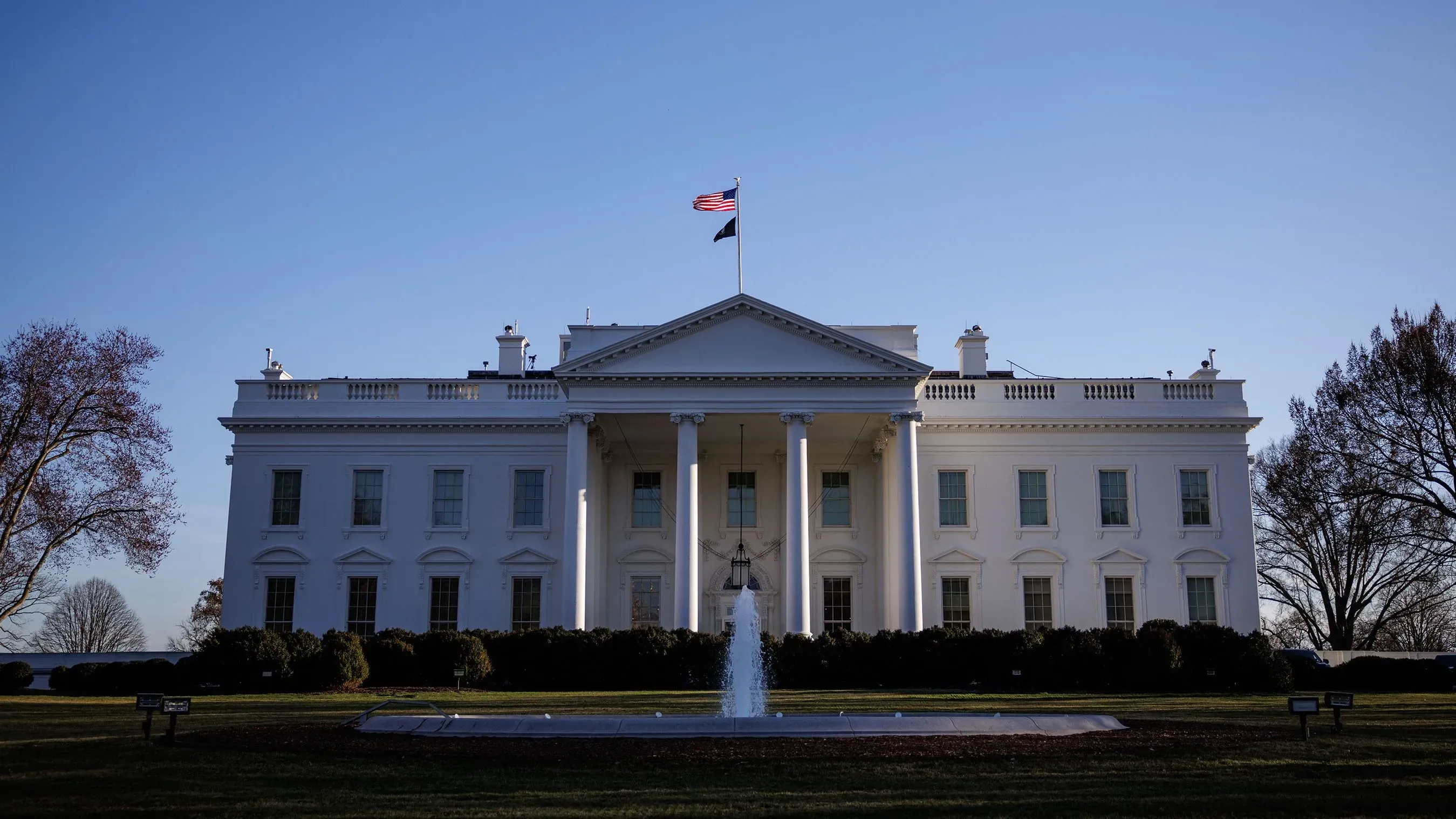 البيت الأبيض مع علم الولايات المتحدة يرفرف في الأعلى، يحيط به حديقة مرتبة، في خلفية سماء زرقاء صافية.