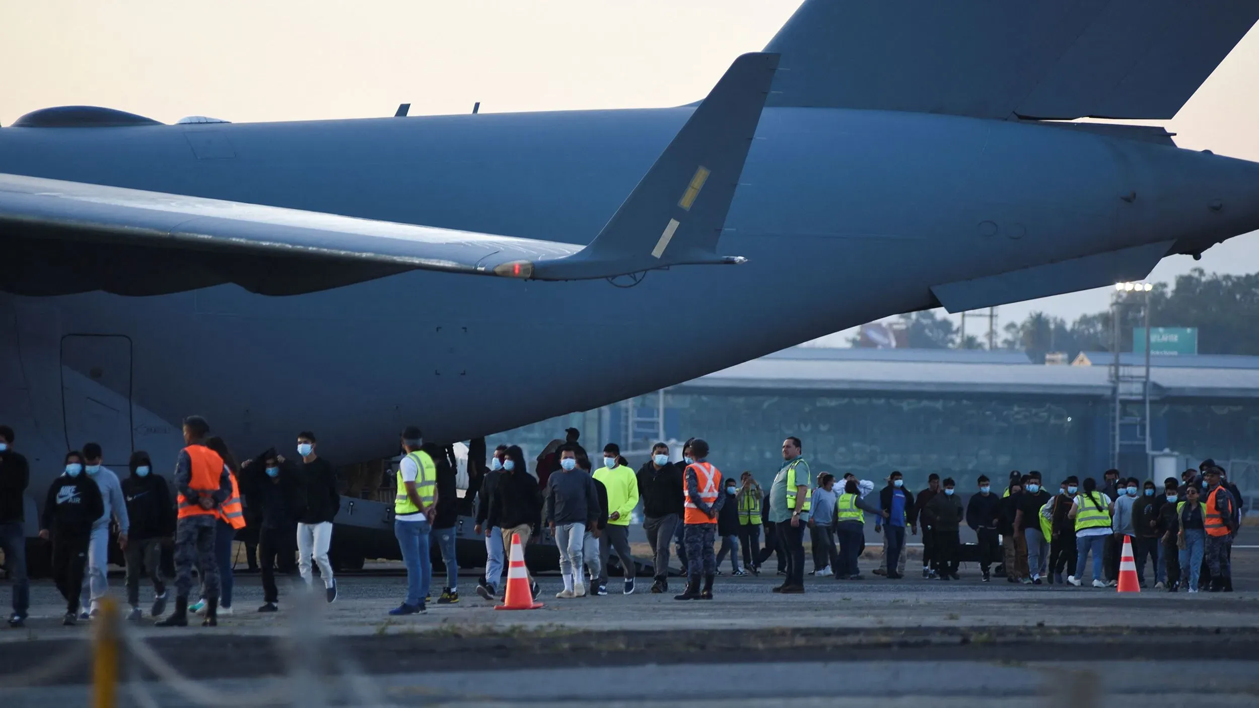 مهاجرون يرتدون أقنعة يتجمعون بالقرب من طائرة شحن عسكرية في موقع ترحيل، وسط إجراءات أمنية مشددة.