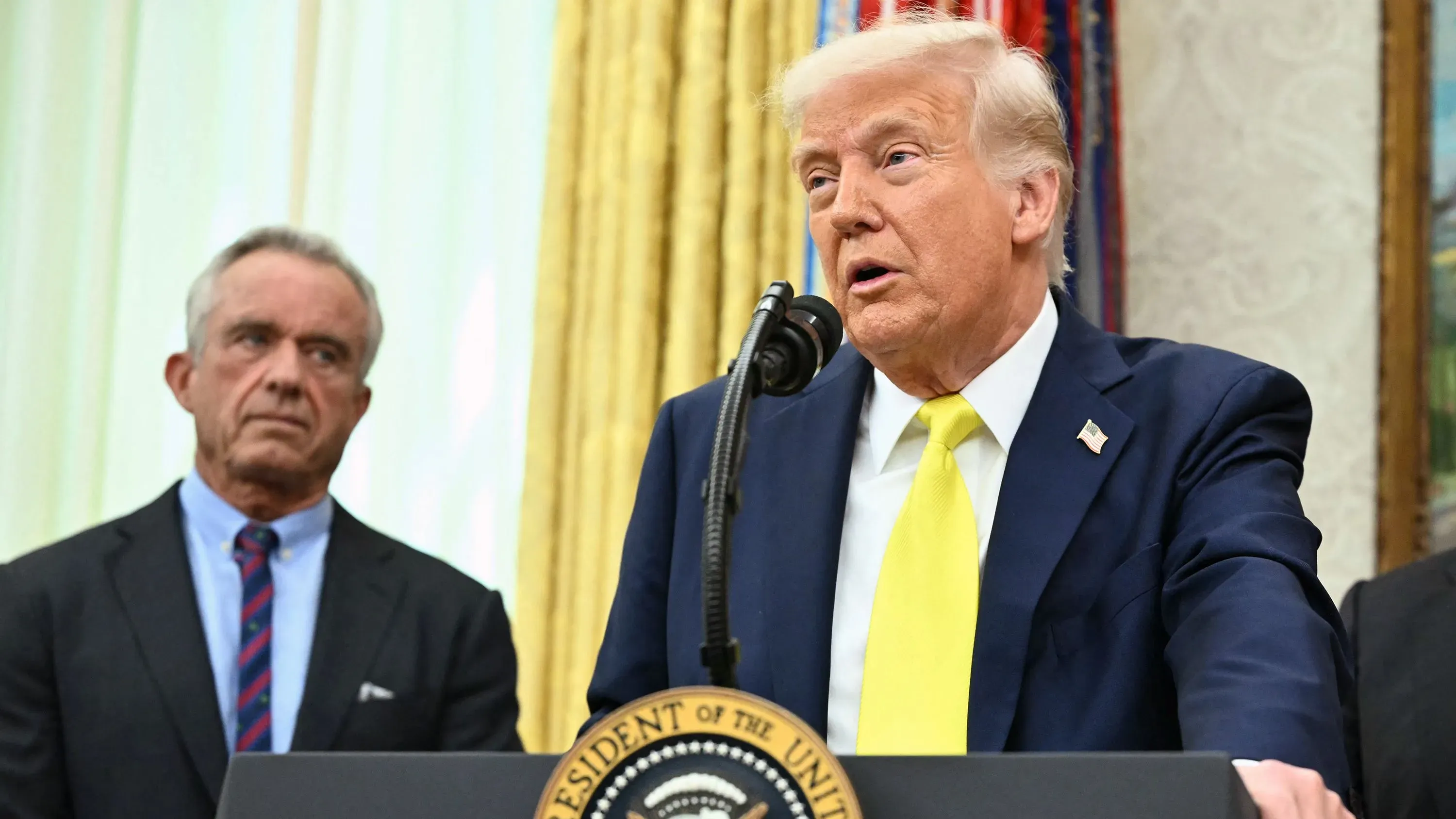 الرئيس السابق دونالد ترامب يتحدث خلال مؤتمر صحفي في المكتب البيضاوي، بينما يظهر روبرت ف. كينيدي جونيور في الخلفية.