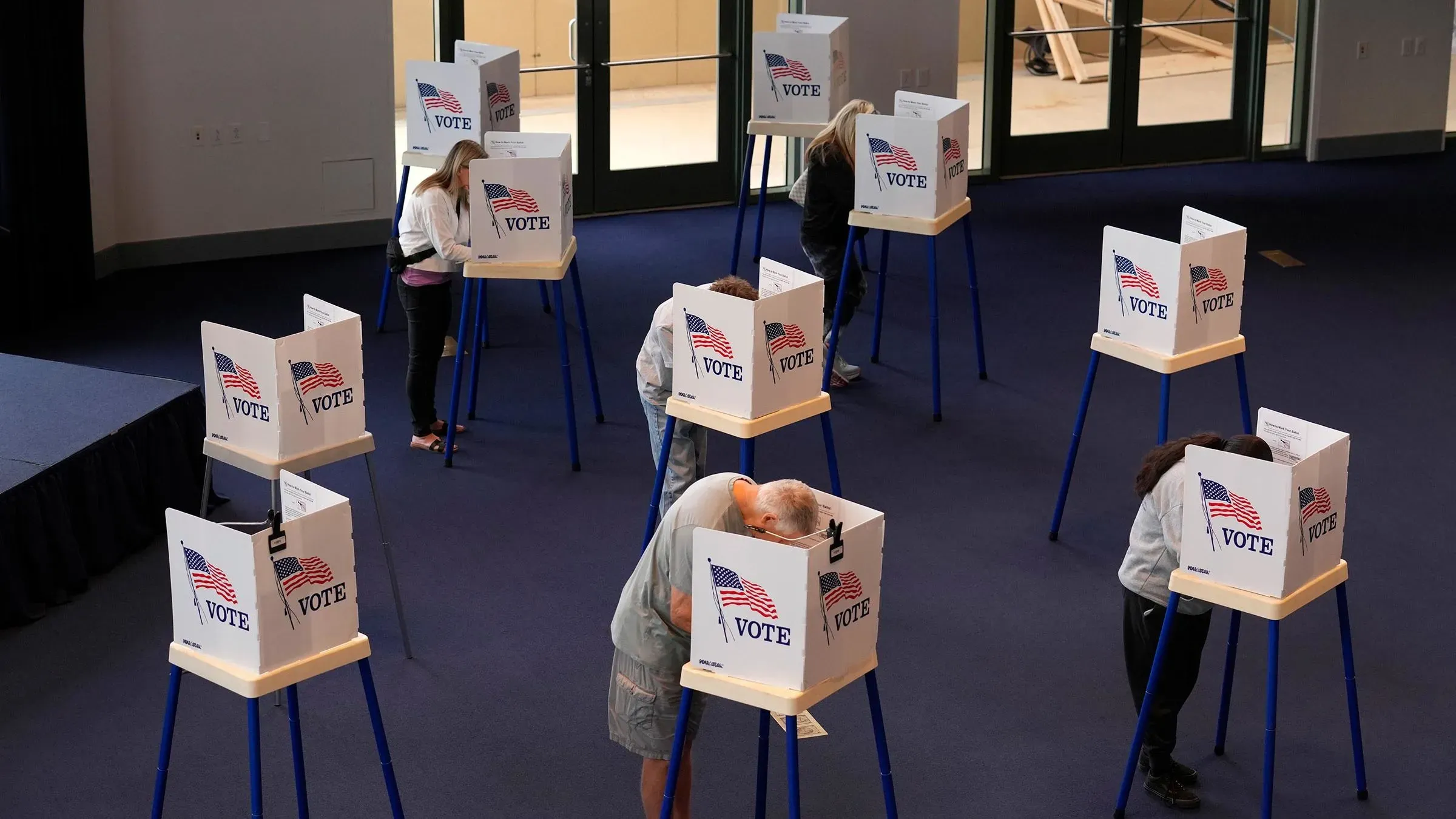 مواطنون يصوتون في مركز انتخابي، حيث تظهر صناديق الاقتراع مع علامات "صوت" وأعلام أمريكية، مما يعكس أهمية الانتخابات في الولايات المتحدة.