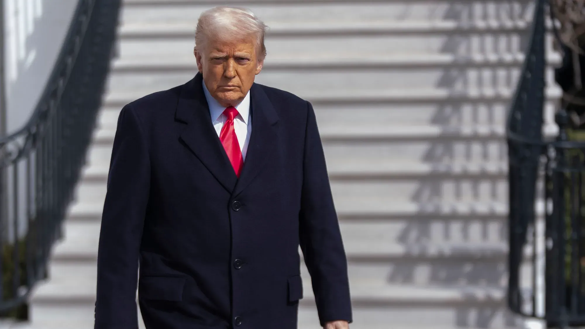 ترامب يسير بخطوات ثابتة خارج البيت الأبيض، مرتديًا معطفًا داكنًا وربطة عنق حمراء، مع تعبير جاد يعكس التوتر الاقتصادي.