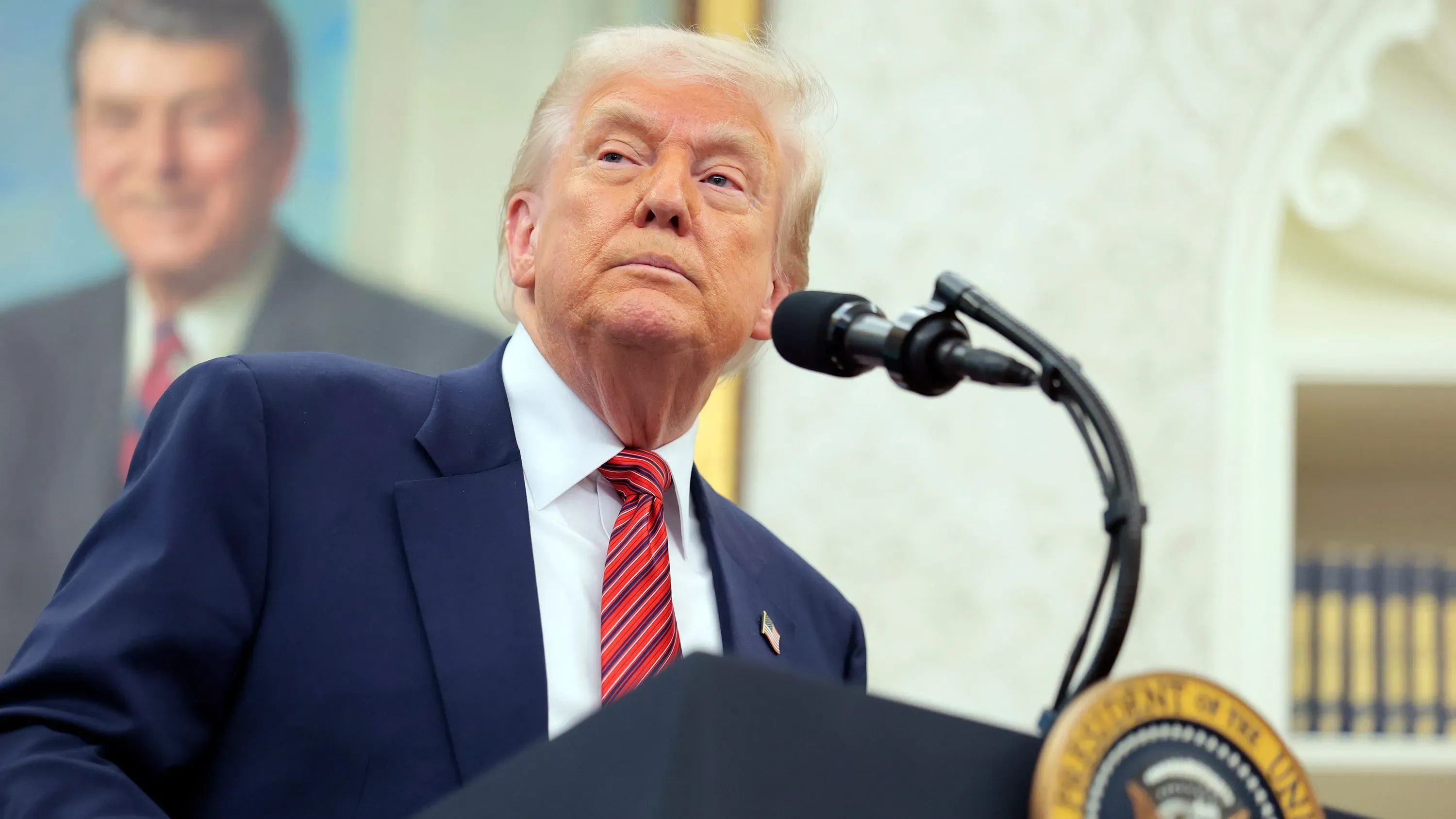 ترامب يتحدث في مؤتمر صحفي، مع صورة سابقة للرئيس رونالد ريغان خلفه، في سياق التوترات التجارية مع الصين وتأثيرها على الاقتصاد الأمريكي.