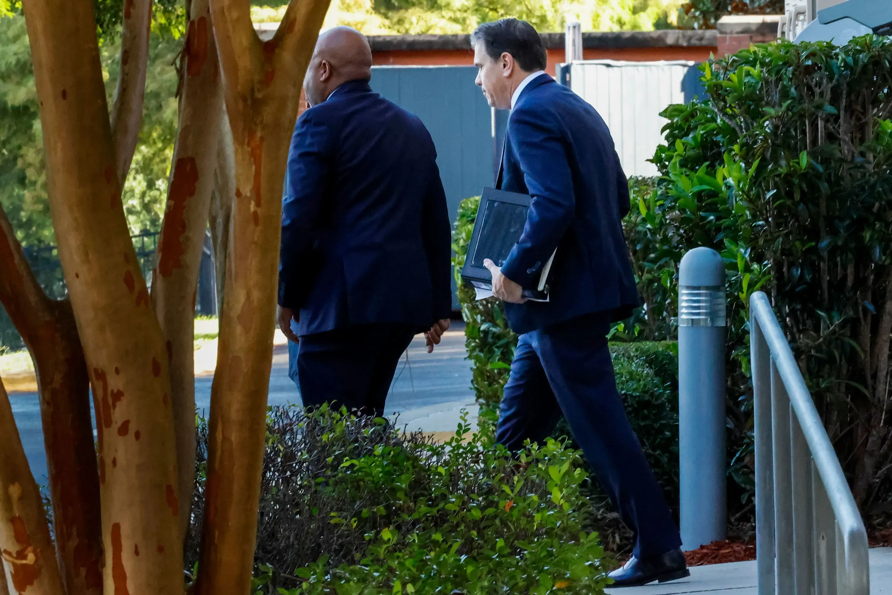 رجلان يرتديان بدلات رسمية يسيران نحو مبنى، أحدهما يحمل ملفًا، بينما يظهر جزء من الأشجار والنباتات المحيطة.