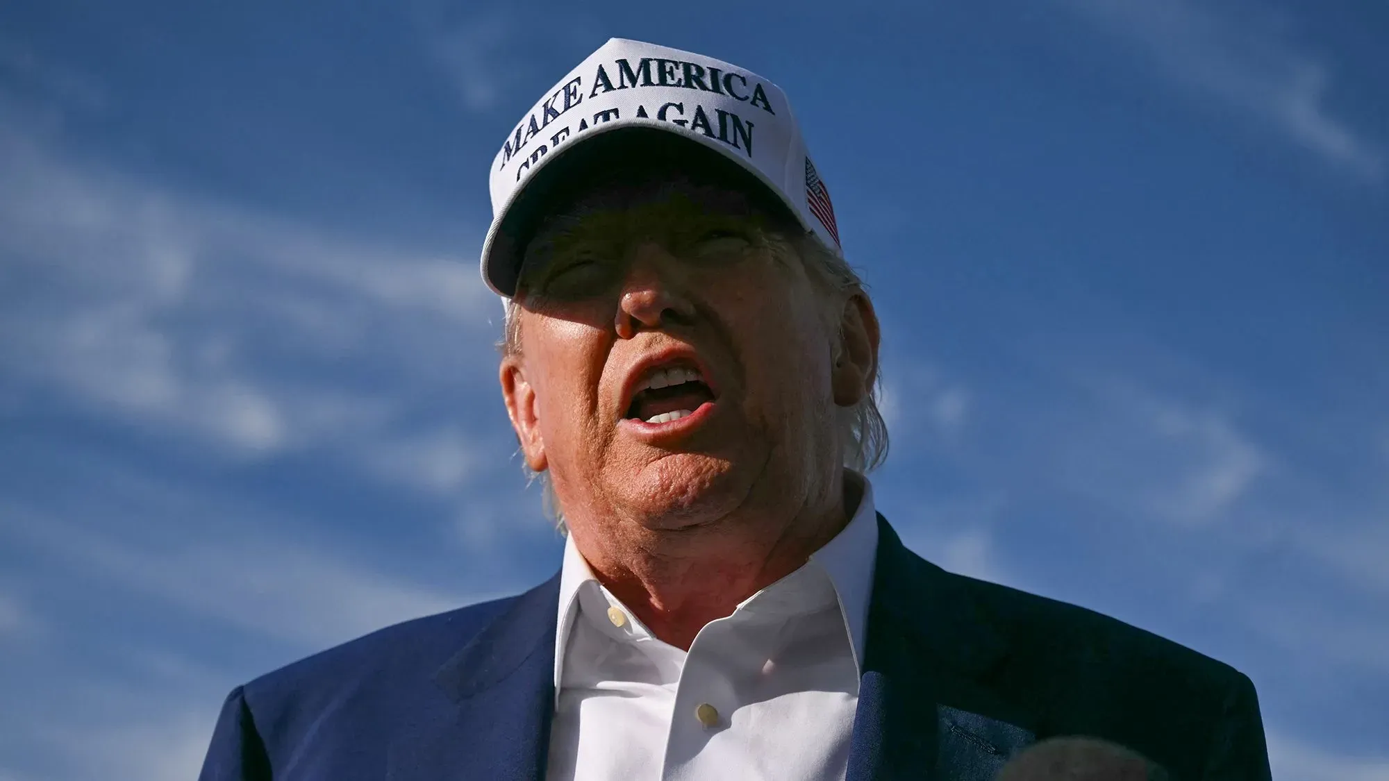 ترامب يتحدث بحدة في مناسبة يوم الذكرى، مرتديًا قبعة تحمل شعار "اجعلوا أمريكا عظيمة مرة أخرى"، مع سماء زرقاء في الخلفية.