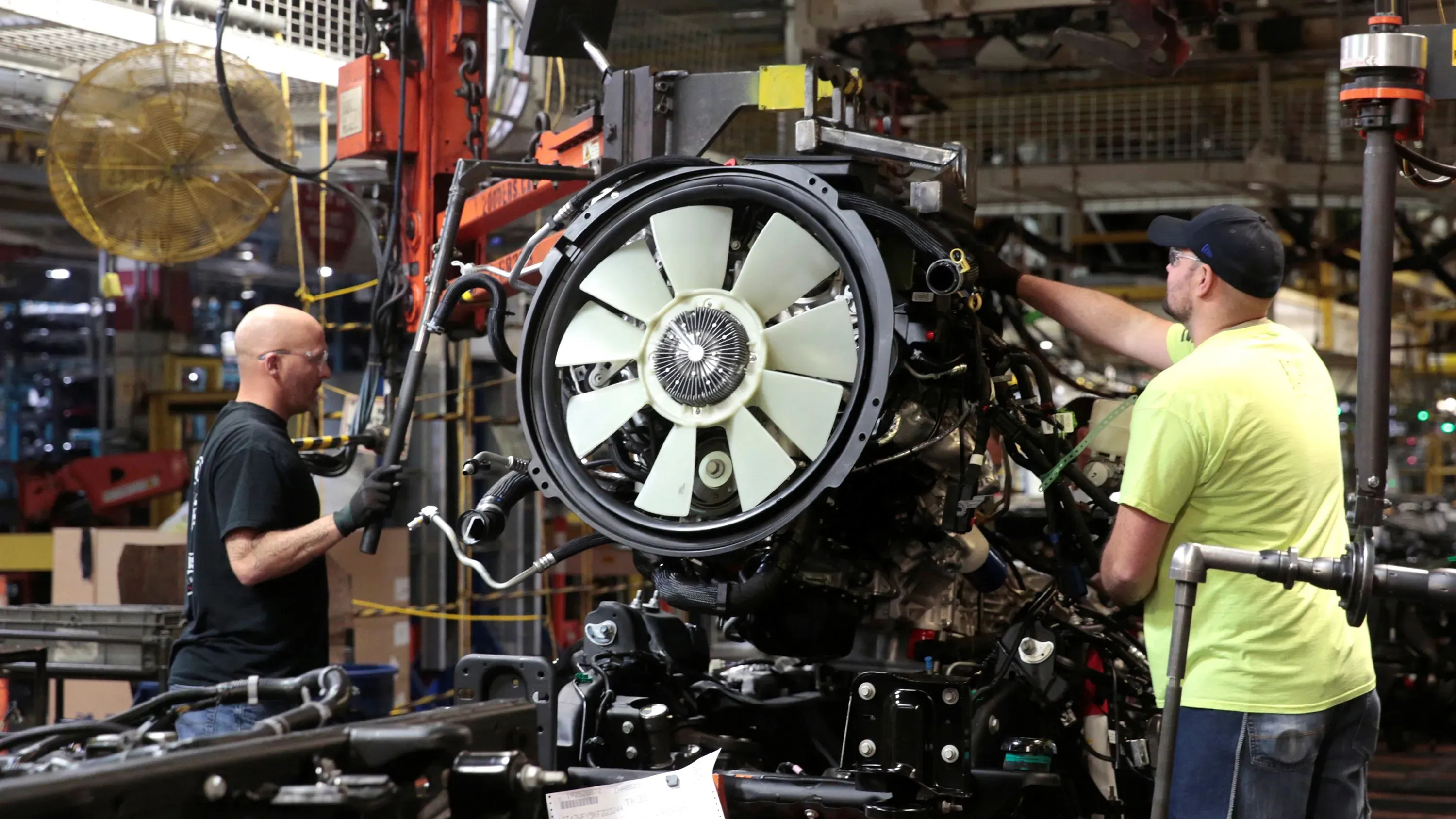 عمال في مصنع سيارات يقومون بتركيب أجزاء المحرك، مع التركيز على التفاصيل الميكانيكية، في سياق النقاش حول تأثير التعريفات الجمركية.