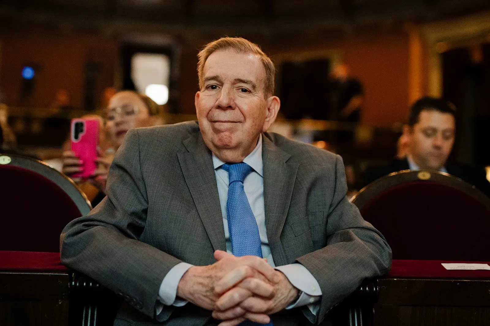 رجل في بدلة رمادية يجلس مبتسمًا في قاعة، مع وجود مشاهدين آخرين في الخلفية. الصورة تعكس أجواء سياسية في سياق الأحداث الفنزويلية.