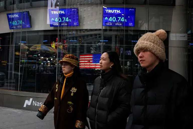 ثلاثة أشخاص يسيرون بجانب واجهة مبنى تعرض شاشات لأسعار أسهم منصة "Truth" مع العلم الأمريكي في الخلفية.