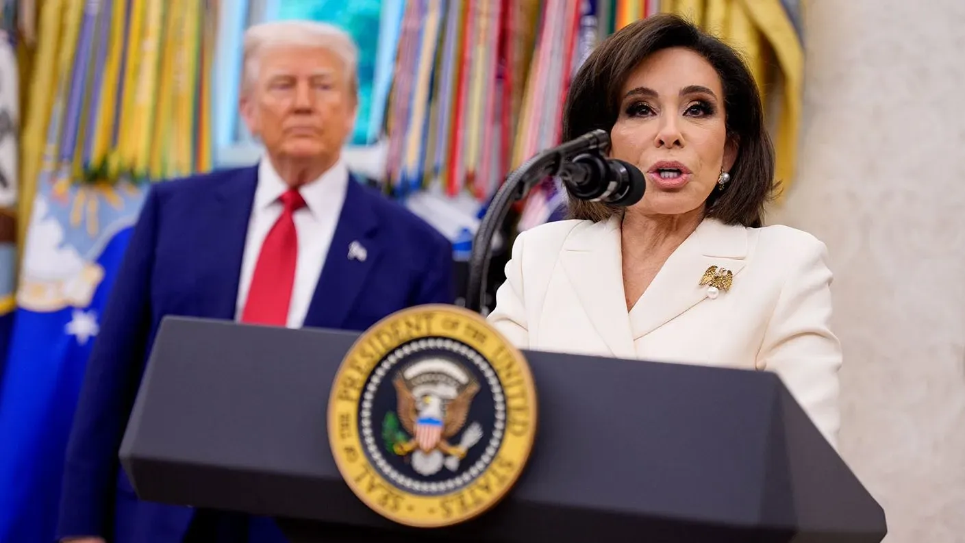 جانين بيرو تتحدث أمام ميكروفون في البيت الأبيض بينما يقف دونالد ترامب خلفها، مع العلم الرئاسي في الخلفية.