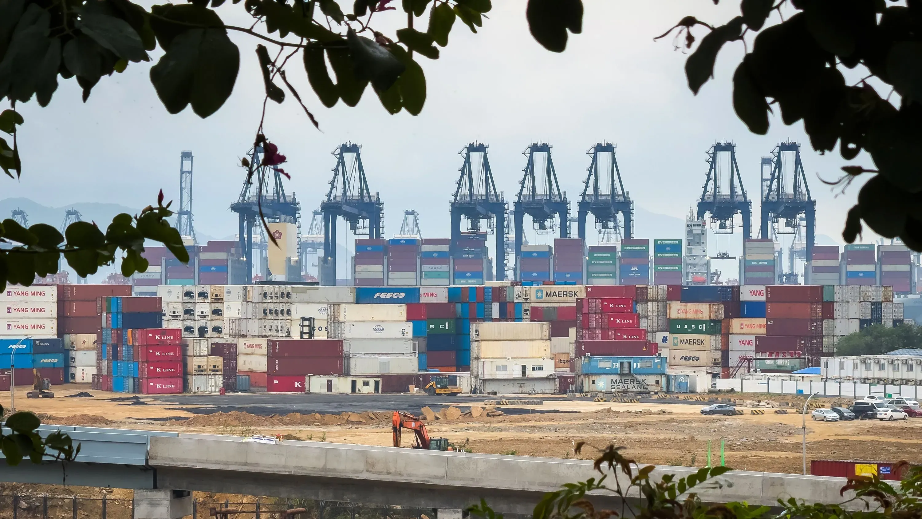 صورة لميناء يحتوي على حاويات شحن متعددة، مع رافعات كبيرة في الخلفية، تعكس تأثير الرسوم الجمركية على التجارة الأمريكية.