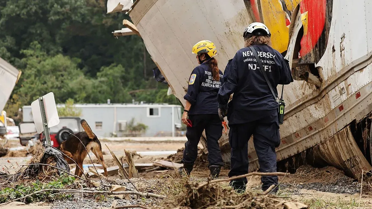 تظهر الصورة فريقًا من رجال الإنقاذ في نيوجيرسي يعملون في موقع دمار بعد كارثة طبيعية، مع وجود كلب بحث يساعدهم في المهمة.