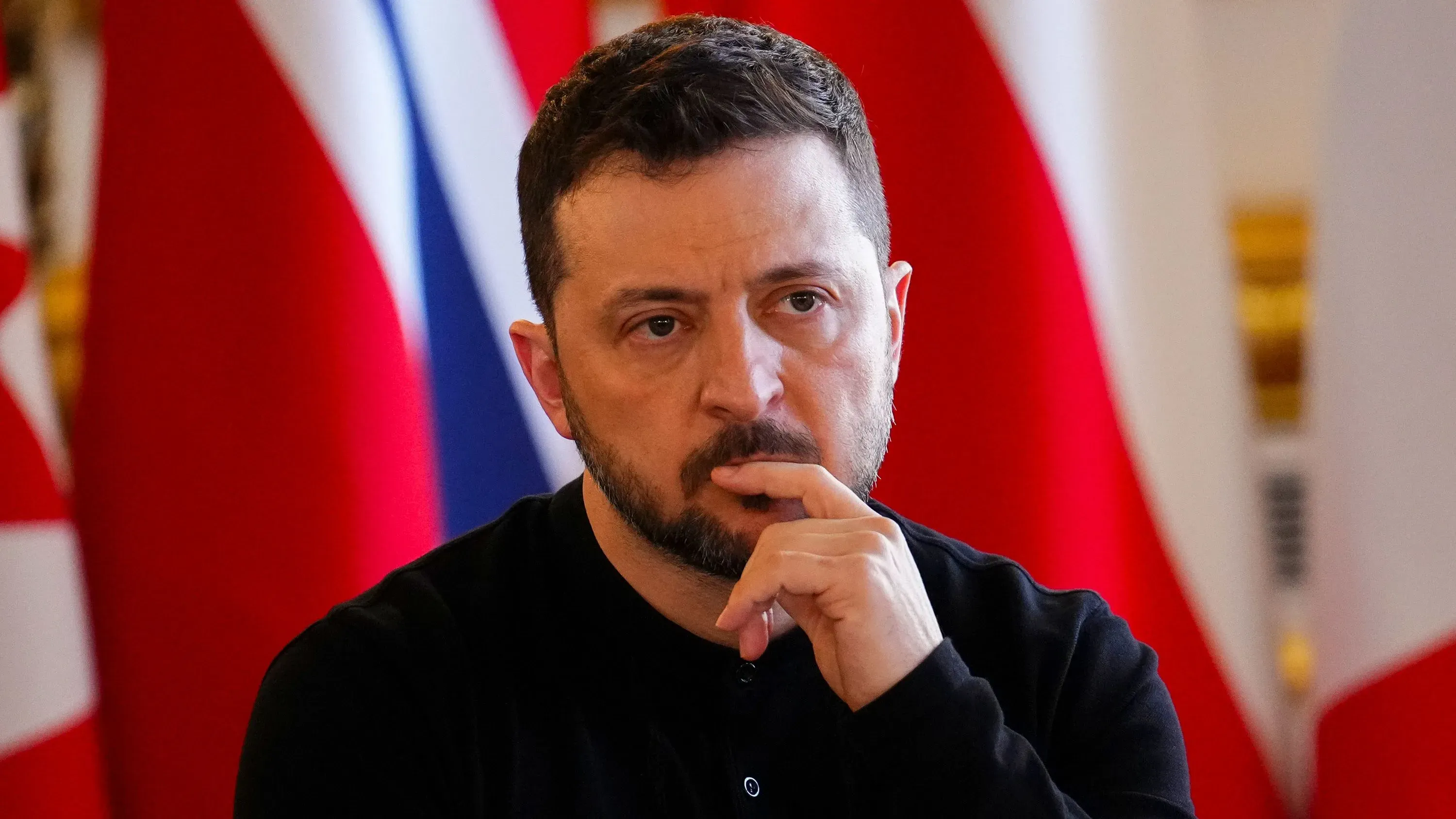 زيلينسكي، الرئيس الأوكراني، يظهر بتعبير جاد خلال مؤتمر صحفي، مع خلفية تضم أعلام دول متعددة، مما يعكس التوترات السياسية الحالية.