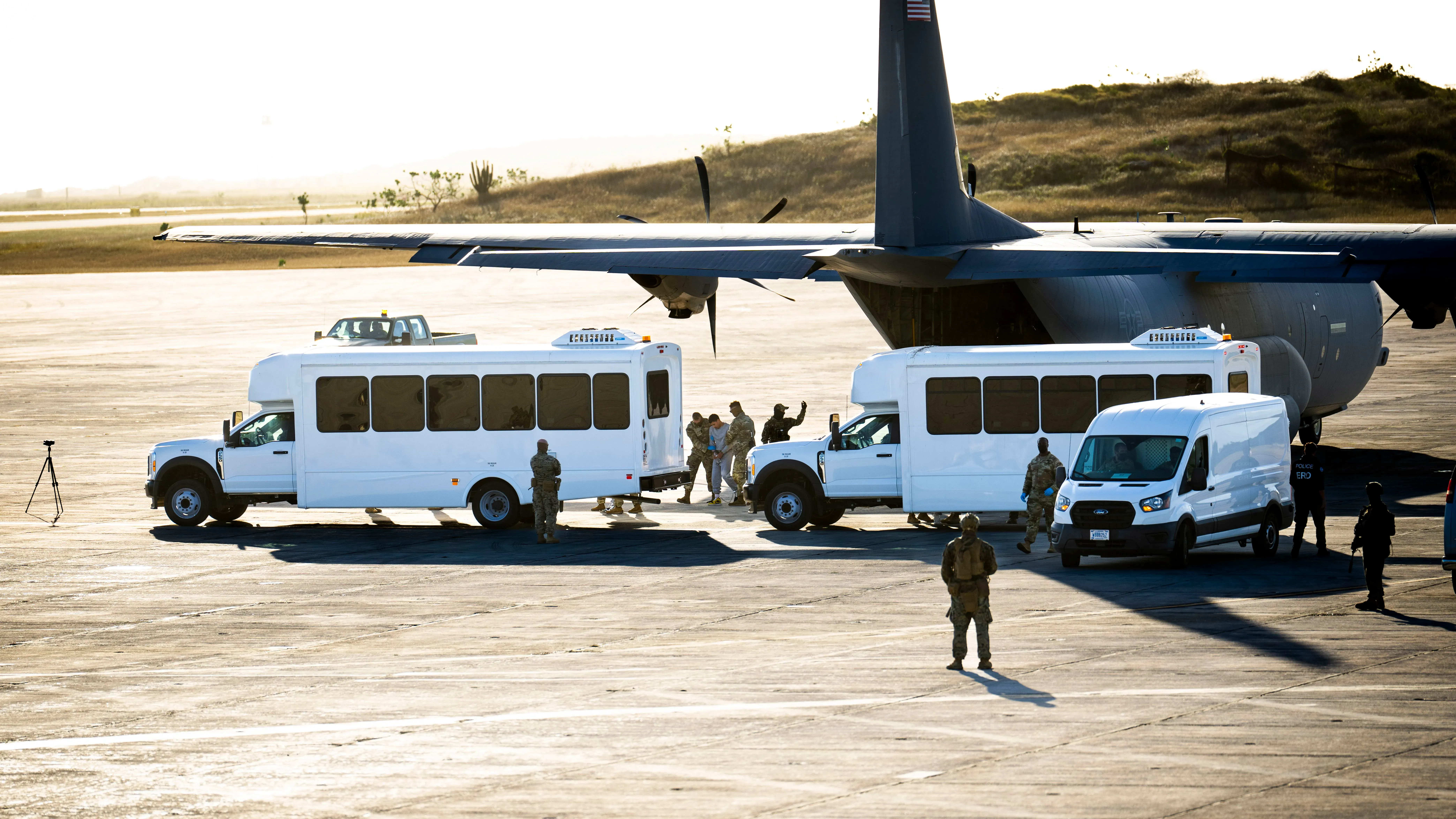 حافلات بيضاء تنتظر على مدرج قاعدة غوانتانامو البحرية، مع وجود جنود أمريكيين، في إطار جهود نقل المهاجرين.