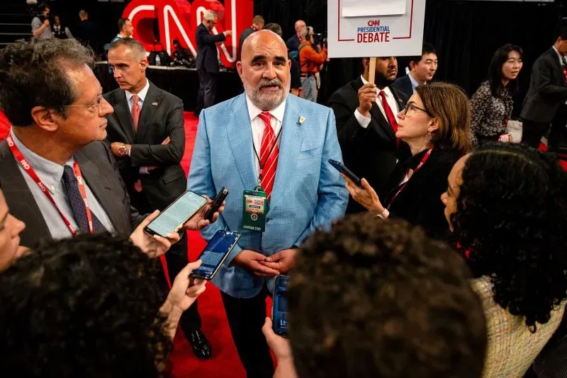 رجل يرتدي بدلة زرقاء يتحدث مع الصحفيين في حدث سياسي، محاطًا بأشخاص يحملون هواتفهم وأعلامًا.