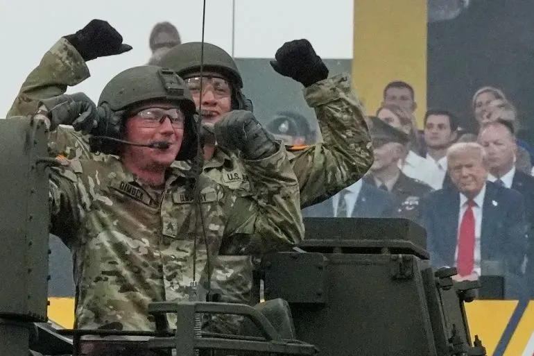 جنود أمريكيون يحتفلون في عرض عسكري بمناسبة عيد ميلاد الجيش الـ250، مع ظهور الرئيس ترامب في الخلفية.