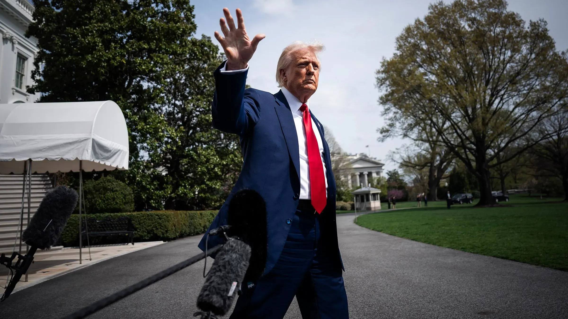 دونالد ترامب يتحدث في حديقة البيت الأبيض، مع ميكروفونات الصحفيين في المقدمة، مع خلفية مبنى حكومي وأشجار.