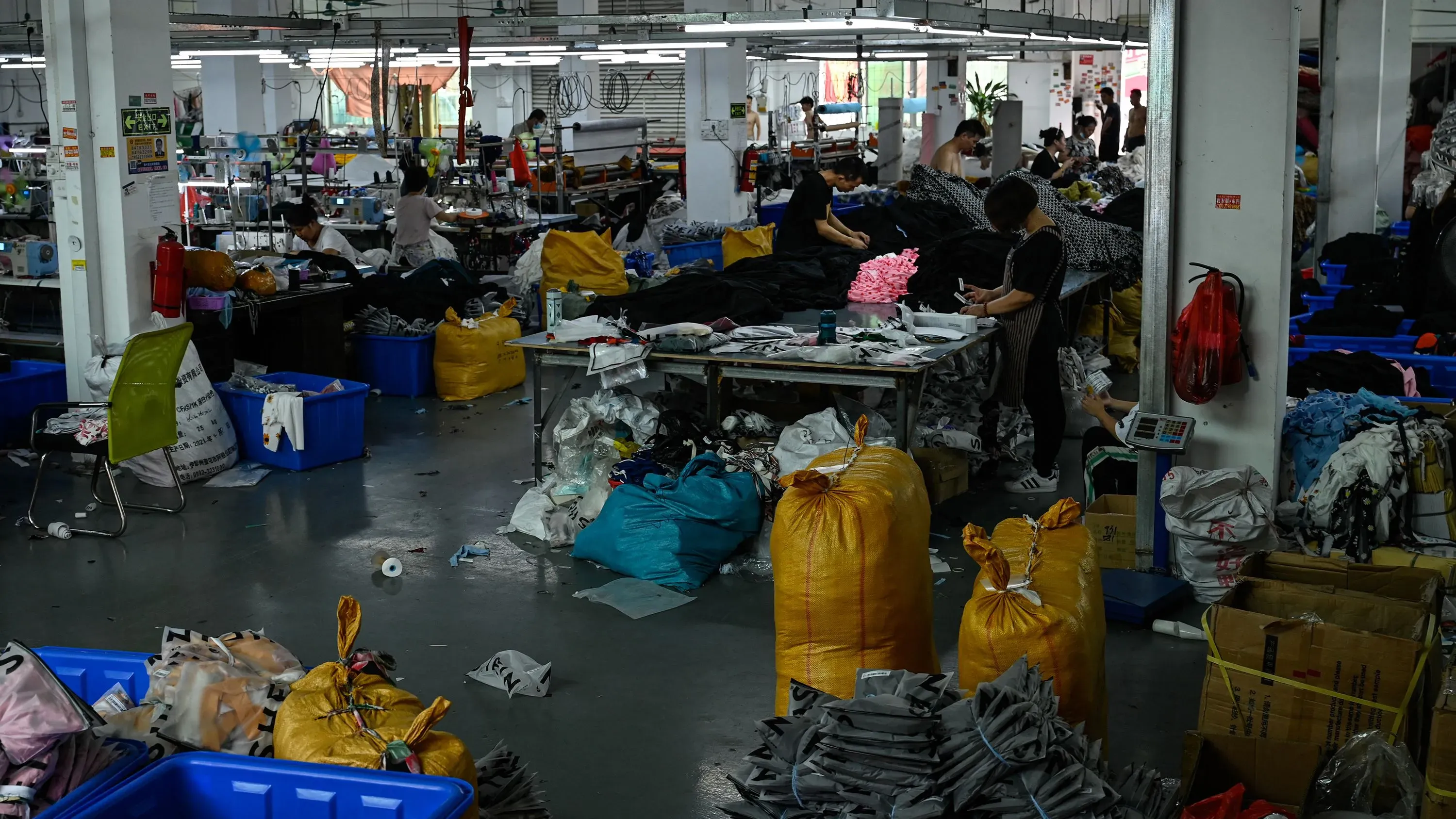 عاملون في مصنع ملابس مزدحم، حيث تُظهر الصورة أكياسًا كبيرة من الأقمشة وأدوات الخياطة، تعكس نشاط التجارة الإلكترونية الصينية.