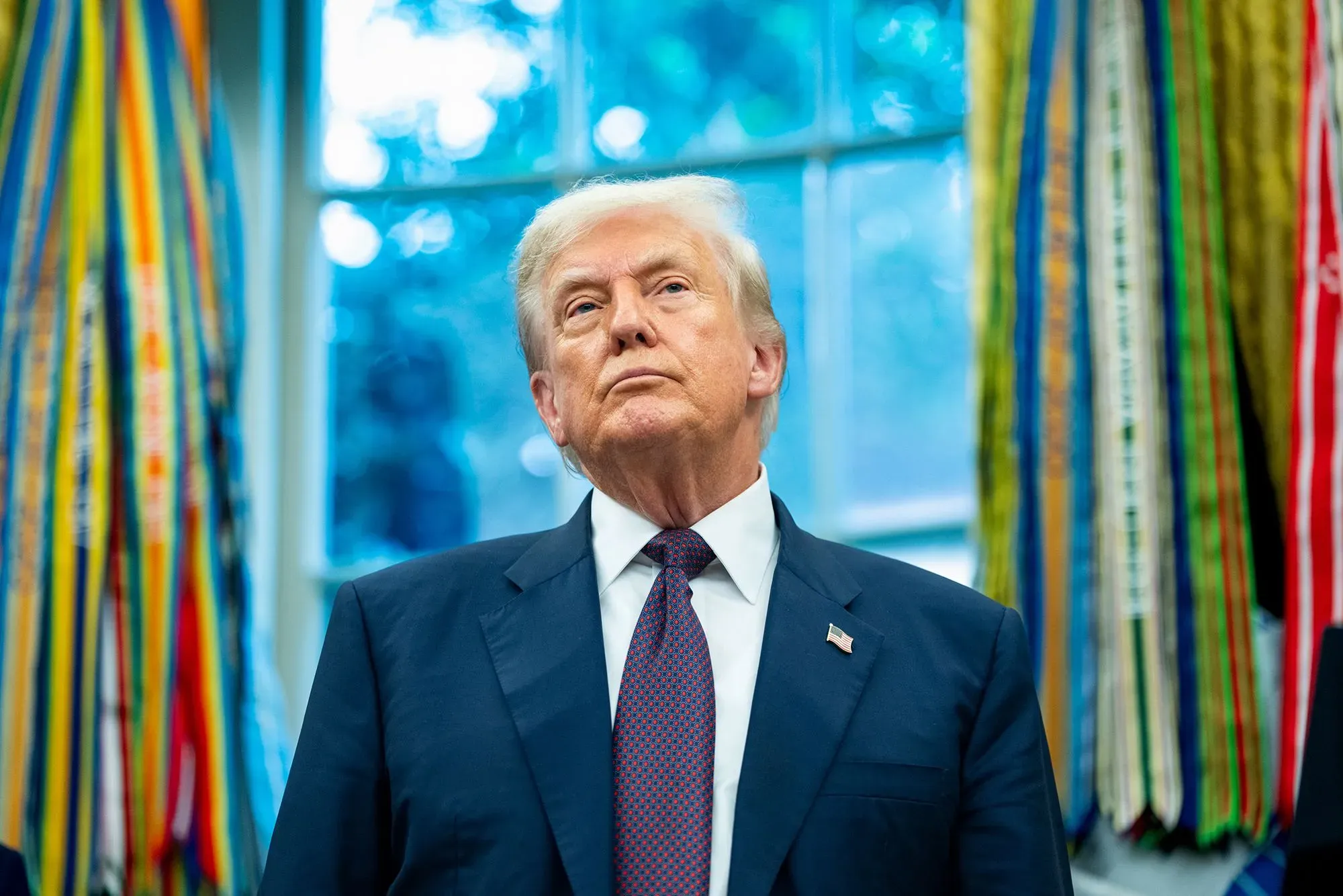 صورة للرئيس الأمريكي دونالد ترامب في المكتب البيضاوي، ينظر إلى الأعلى بتعبير جاد، محاطًا بأشرطة ملونة تعكس تأثيره على النظام التجاري العالمي.