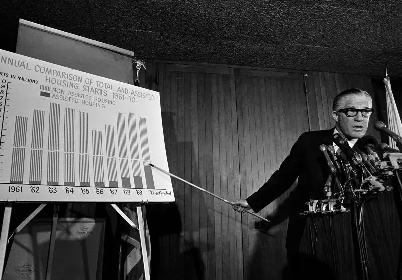 رجل يتحدث أمام مجموعة من الميكروفونات، مشيرًا إلى رسم بياني يقارن بين الإسكان المدعوم وغير المدعوم في الفترة من 1961 إلى 1970.