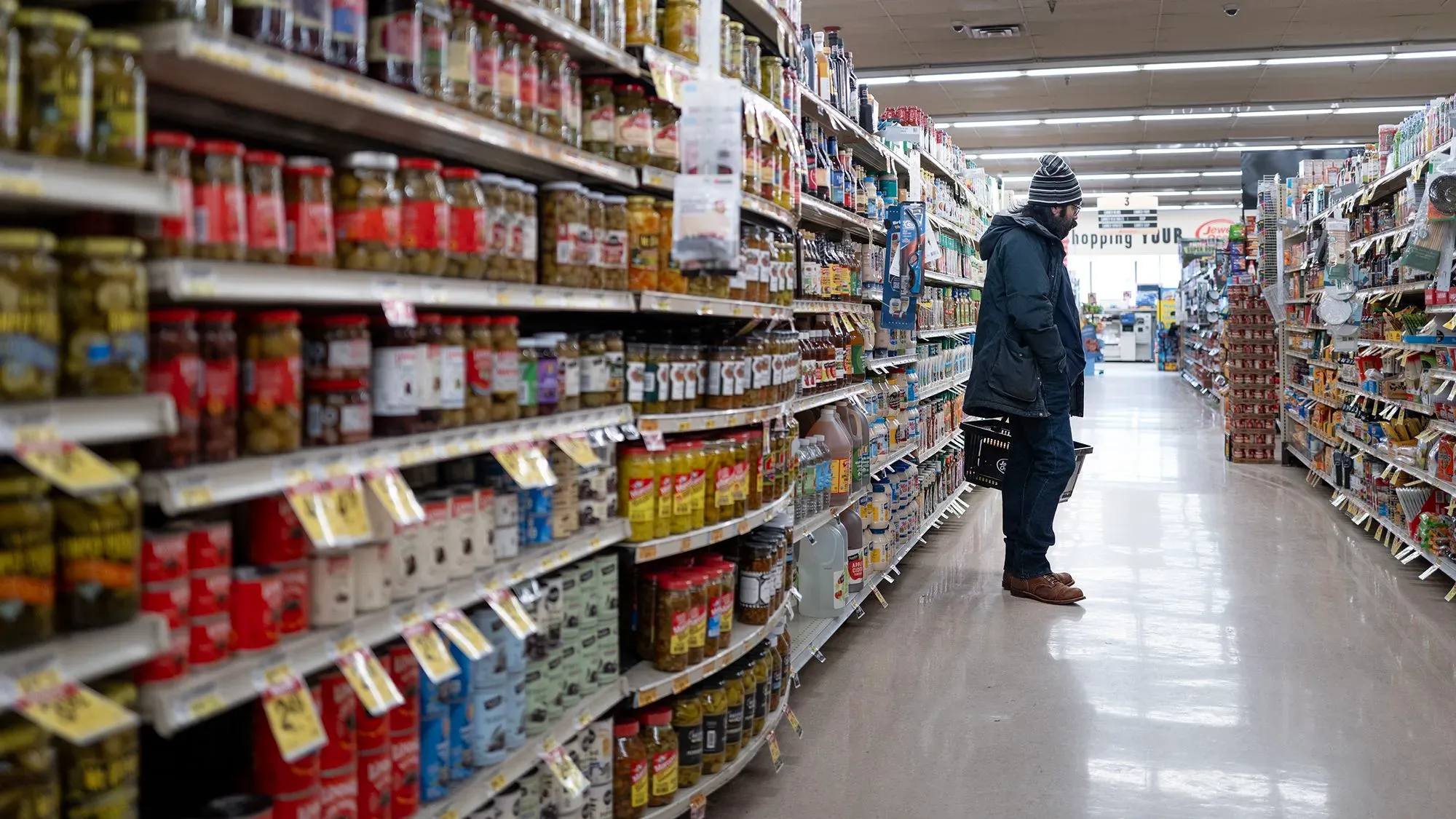 رجل يقف في ممر سوبرماركت ويحمل سلة تسوق، محاط بمختلف الأطعمة المعلبة والمنتجات، في سياق أزمة أسعار المواد الغذائية.