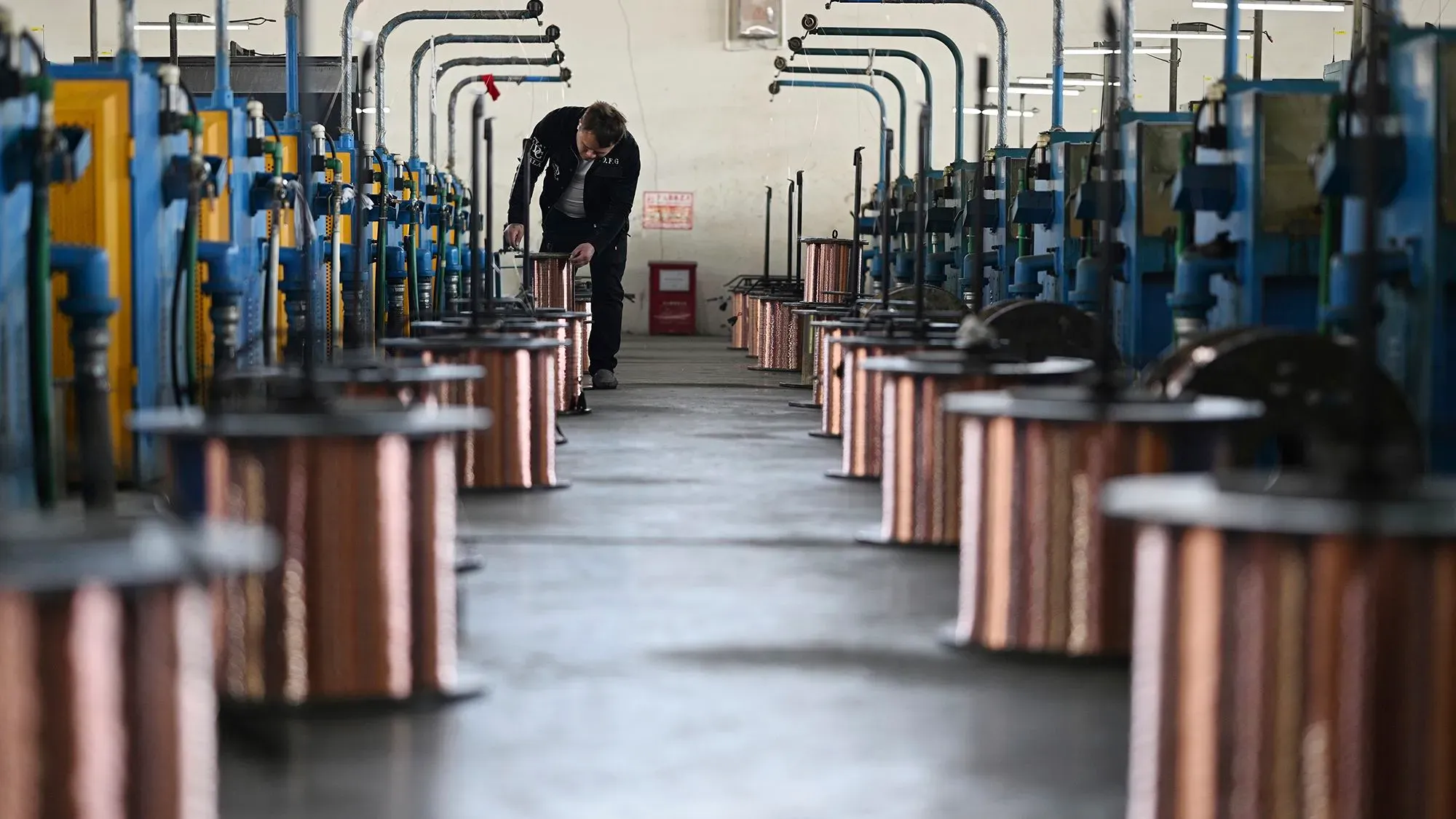 عامل في مصنع للنحاس يتفقد أسلاك النحاس الملفوفة، مع وجود آلات تصنيع في الخلفية، في سياق تأثير التعريفات الجمركية.
