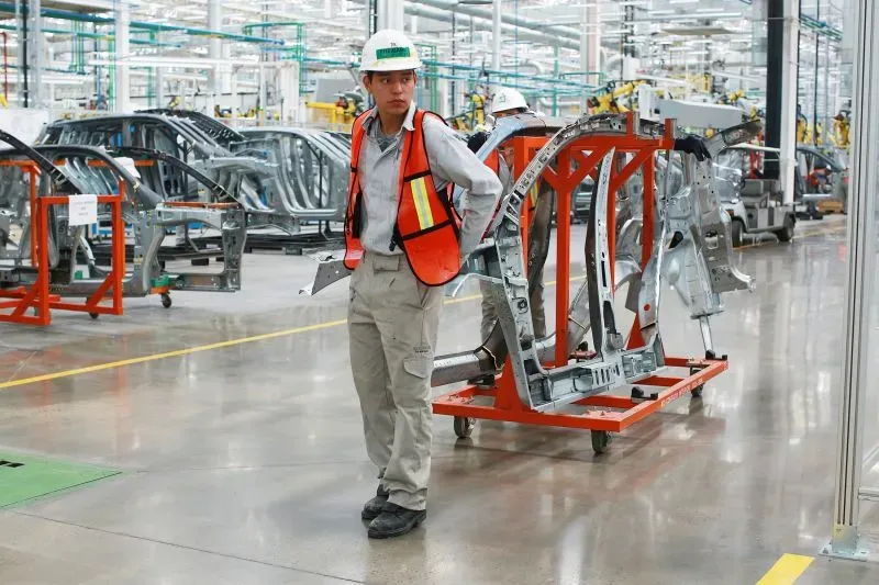 عامل في مصنع سيارات يحمل هيكل سيارة على عربة، مع معدات تصنيع حديثة في الخلفية، مما يعكس تأثير الأتمتة على صناعة السيارات الأمريكية.