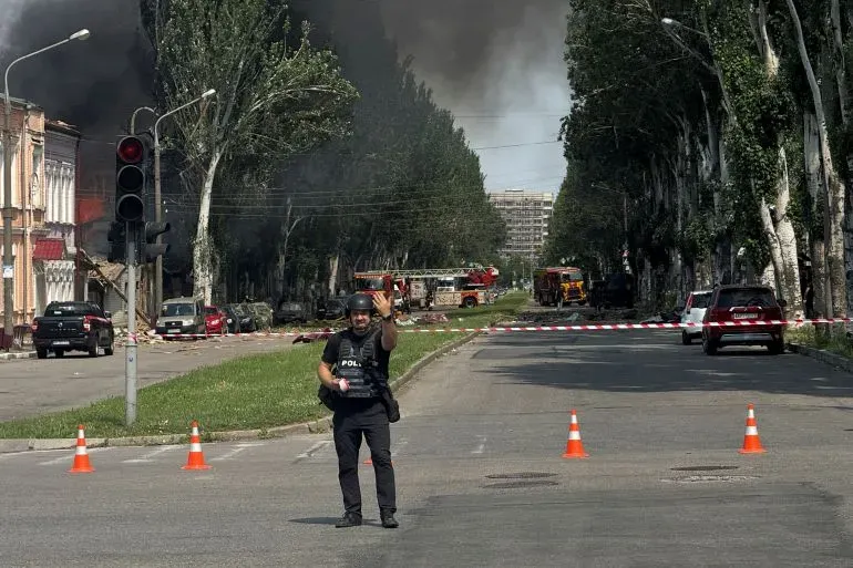 شرطي يرتدي خوذة ودوريات في موقع حريق بمدينة أوكرانية، مع وجود سيارات إسعاف وشرطة في الخلفية وسط دخان كثيف.