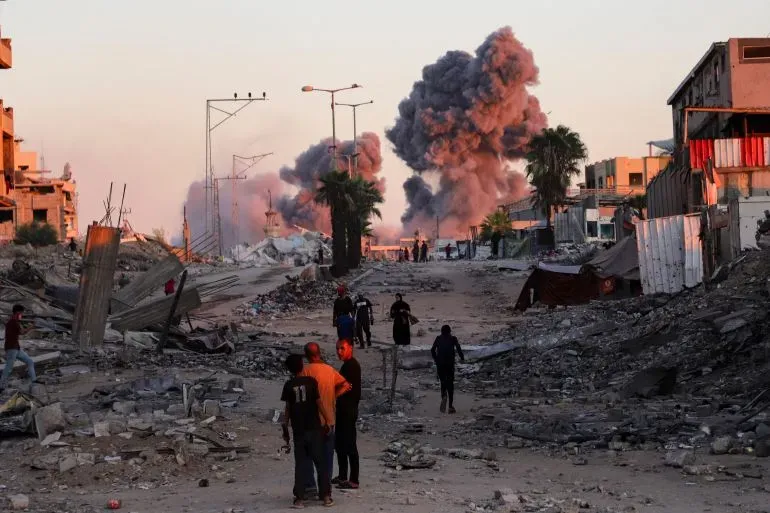 دخان يتصاعد من انفجارات في غزة، مع وجود أشخاص يسيرون بين الأنقاض والمباني المدمرة، مما يعكس تأثير الحرب المستمر على المدنيين.