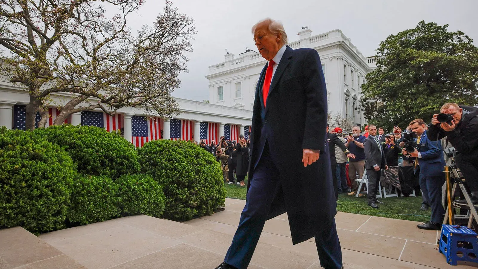 الرئيس ترامب يسير في حديقة البيت الأبيض، محاطًا بالمراسلين، بينما تبرز الأعلام الأمريكية خلفه، في أجواء من التوتر السياسي.