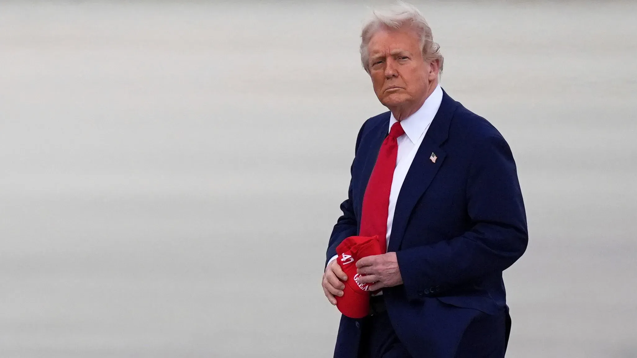 الرئيس الأمريكي دونالد ترامب يرتدي بدلة زرقاء وقميصًا أبيض، ممسكًا بقبعة حمراء، أثناء مغادرته مكان ما.