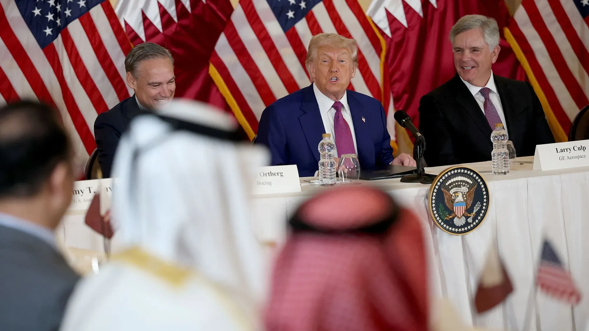 اجتماع مائدة مستديرة في قطر حيث يتحدث ترامب عن إلغاء الرسوم الجمركية مع قادة الأعمال، مع وجود أعلام الولايات المتحدة وقطر خلفهم.