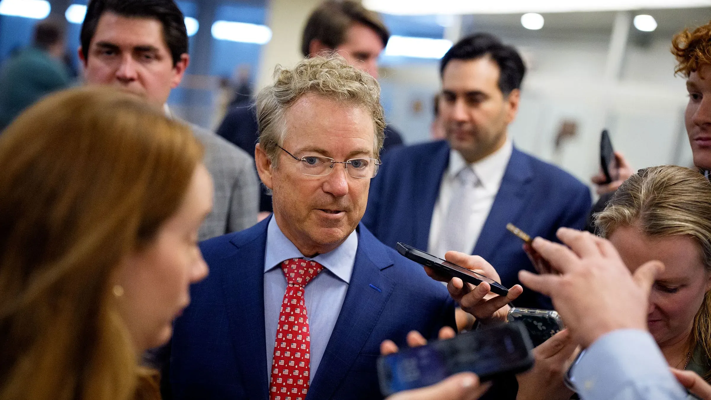السناتور راند بول يتحدث مع الصحفيين وسط حشد من المراسلين، مع التركيز على قضايا قانونية تتعلق بالسياسة المالية.