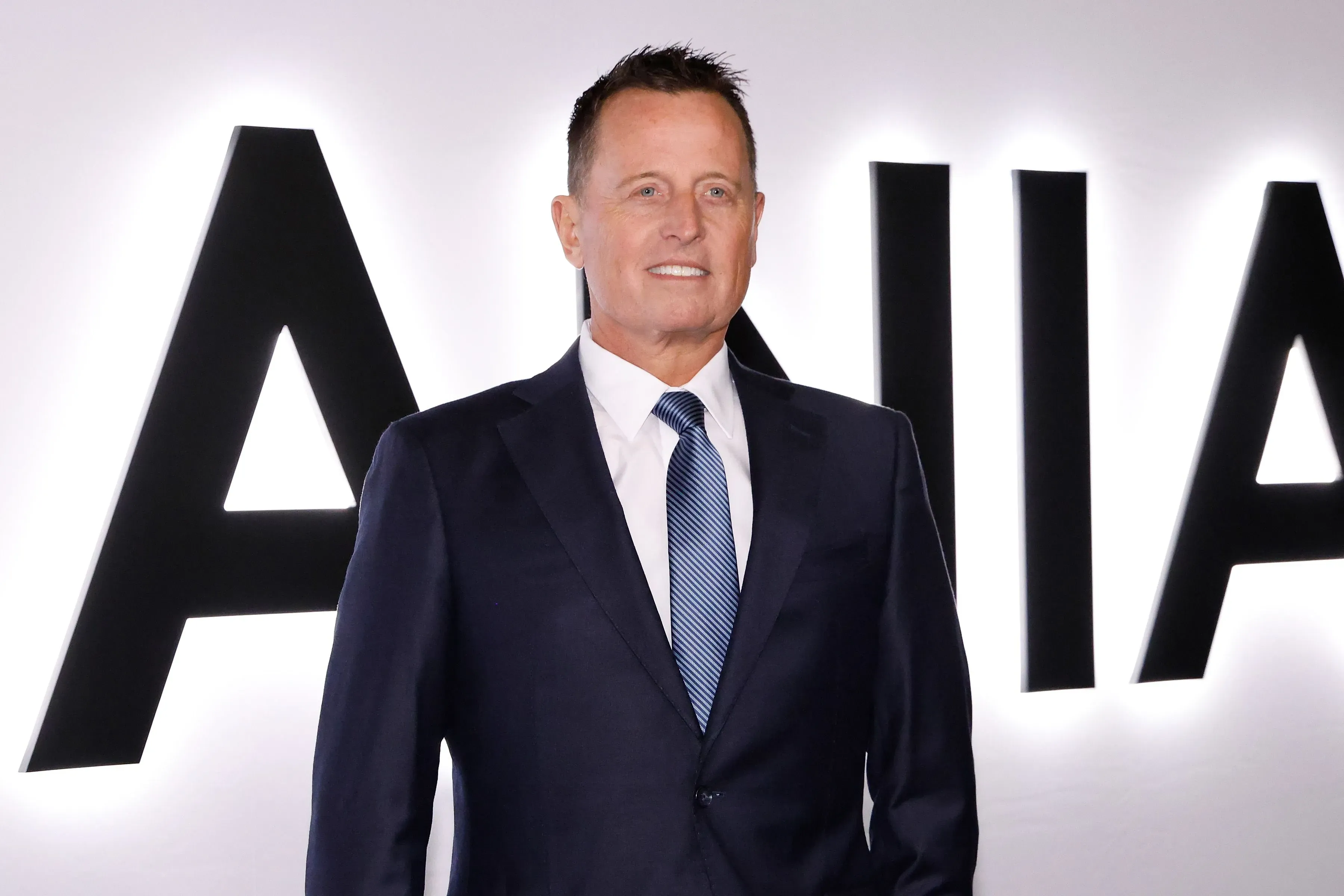 ريتشارد غرينيل، الرئيس السابق لمركز كينيدي، يظهر مبتسمًا في بدلة رسمية أمام شعار المؤسسة، وسط أجواء من التوتر والاحتجاجات.