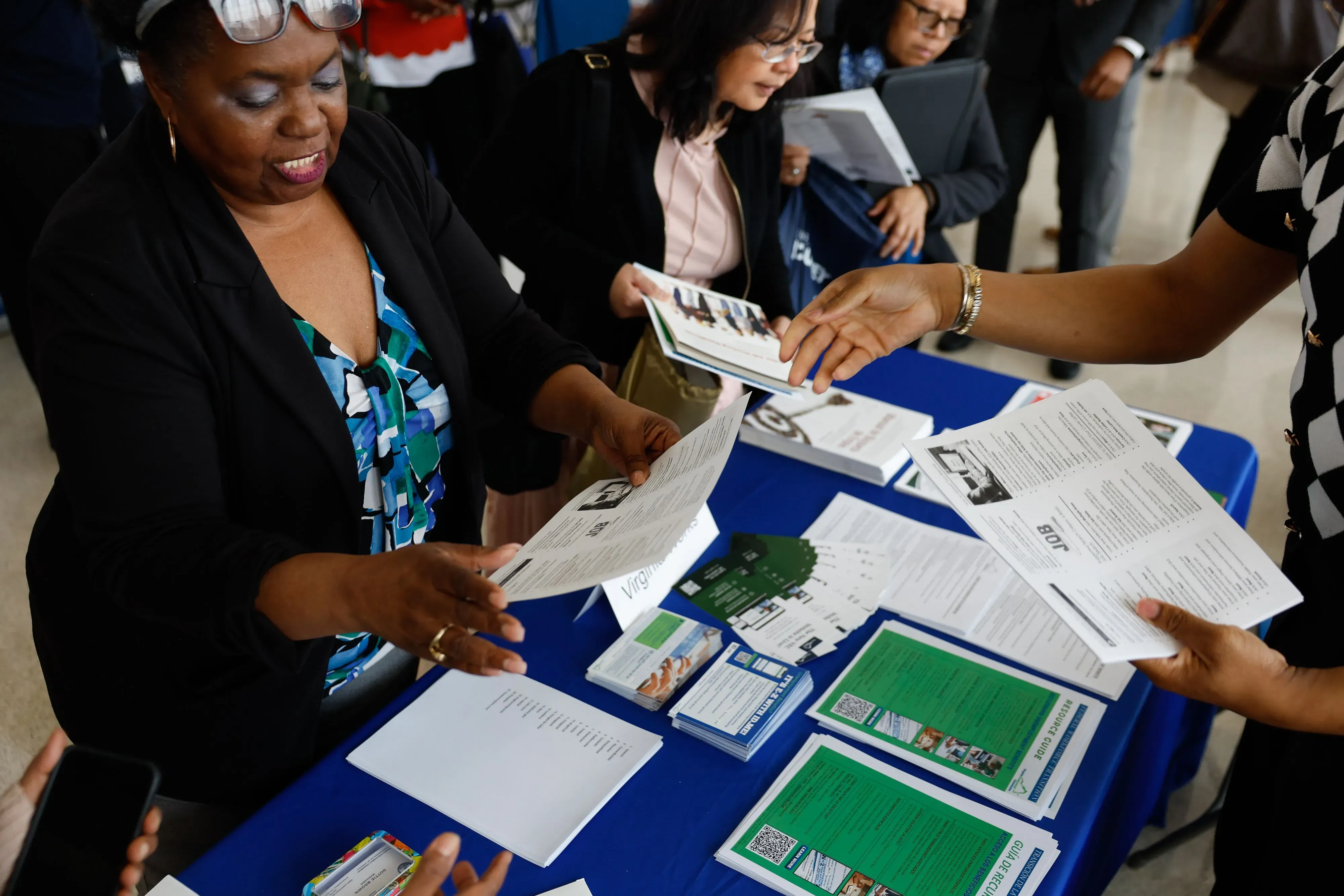 امرأة تتفاعل مع شخص آخر في حدث توظيف، حيث تتبادل منشورات ومعلومات حول فرص العمل. خلفية تحتوي على مجموعة من الحضور.