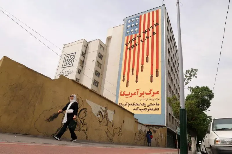 امرأة ترتدي حجابًا تسير بجوار جدار يحمل لوحة جدارية كبيرة تظهر علم الولايات المتحدة مع صواريخ، تعبر عن مشاعر معارضة تجاه أمريكا.