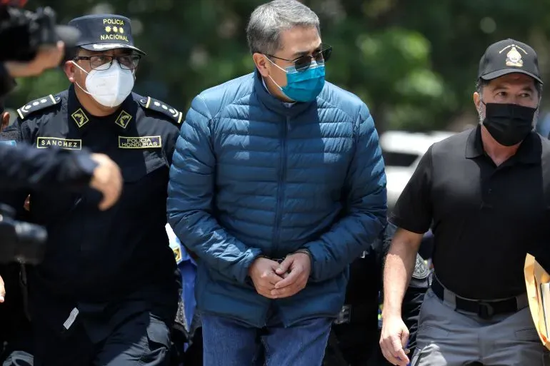 رئيس هندوراس السابق خوان أورلاندو هيرنانديز محاطاً برجال الشرطة، مع التركيز على ملابسه الزرقاء وقناع الوجه، في سياق قضايا الاتجار بالمخدرات.