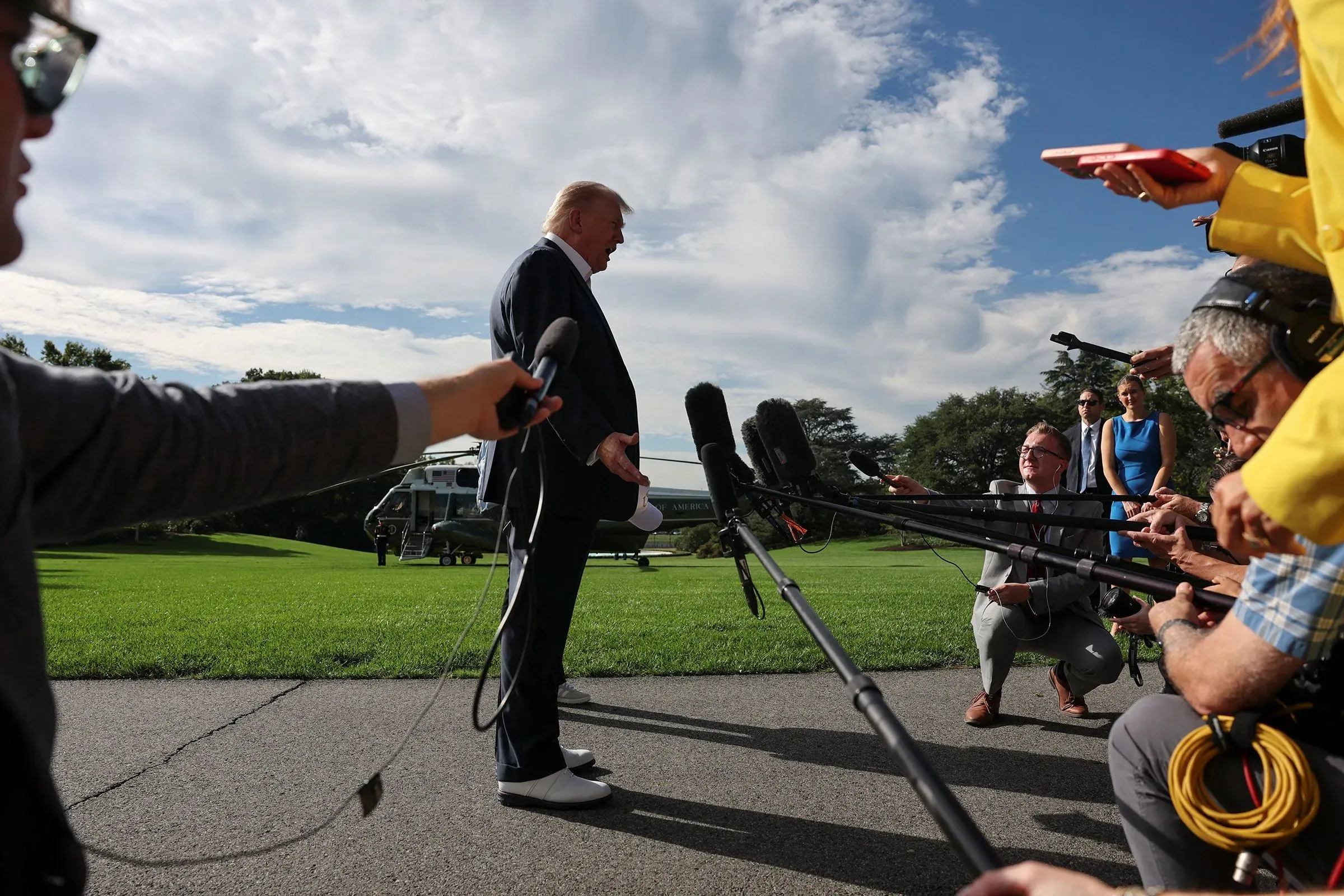 ترامب يتحدث إلى الصحفيين مع مجموعة من الميكروفونات، بينما يظهر في الخلف طائرة هليكوبتر. الجو مشمس والخلفية خضراء.