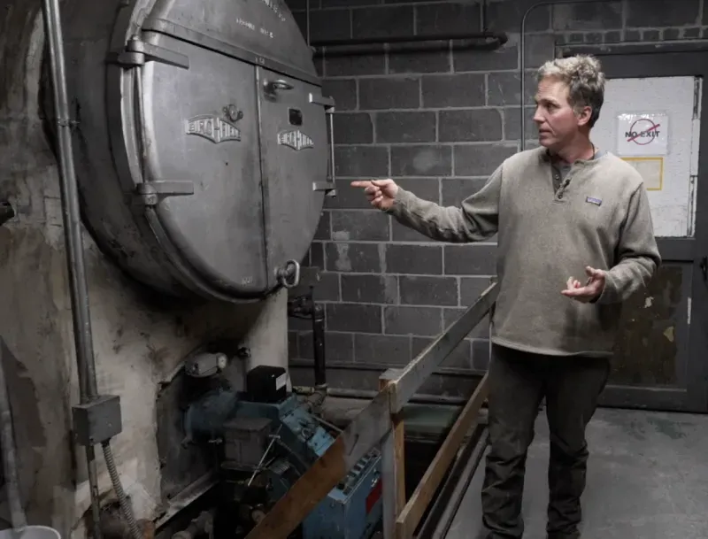 رجل يتحدث بجوار غرفة ضاغطة قديمة في مكان صناعي، مع تفاصيل عن المعدات المحيطة، تعكس موضوع الفساد الحكومي وتأثيره.