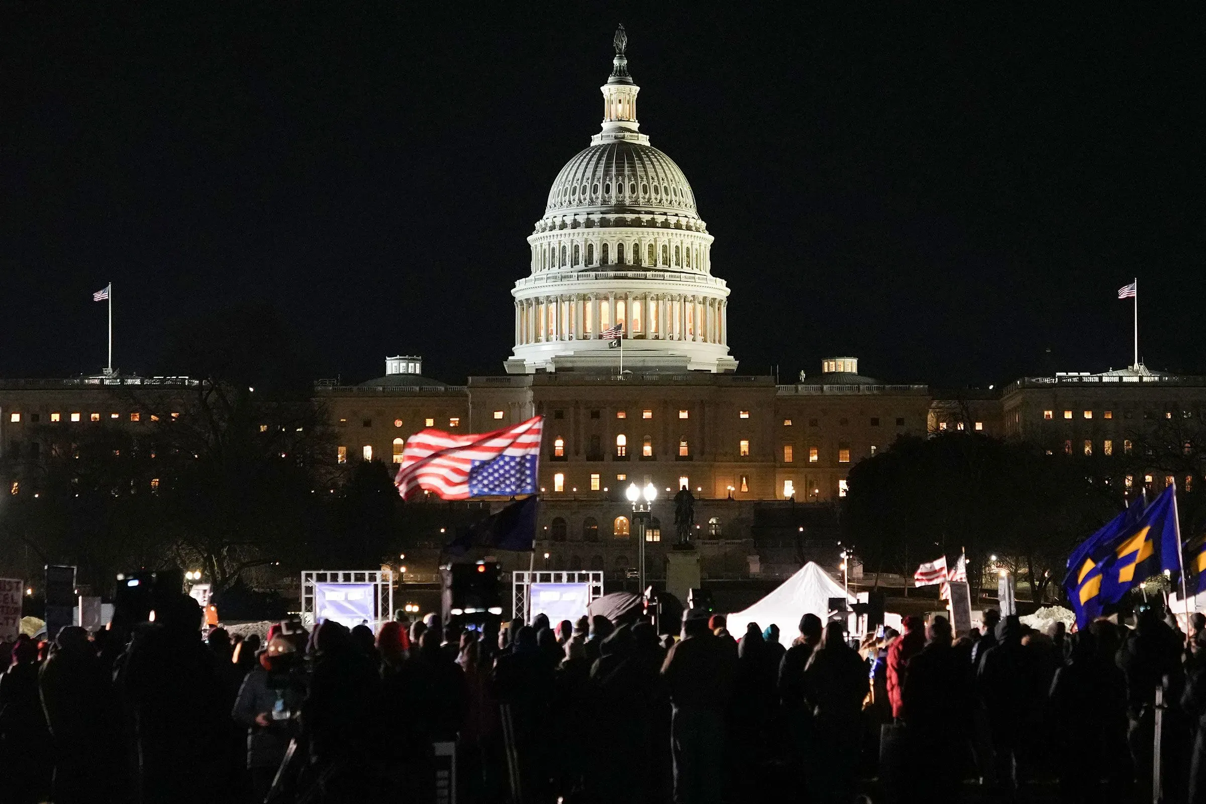 مظاهرة حاشدة أمام مبنى الكابيتول الأمريكي في الليل، مع أعلام ترفرف في الهواء وأضواء المبنى تضيء المشهد.